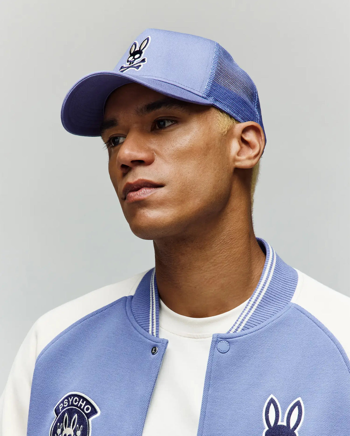 A person with short blond hair wears the light blue Psycho Bunny MENS RYDER TRUCKER CAP (MO1500030), an adjustable snapback, and a matching varsity jacket with a bunny logo, gazing slightly upward and to the side against a plain background.