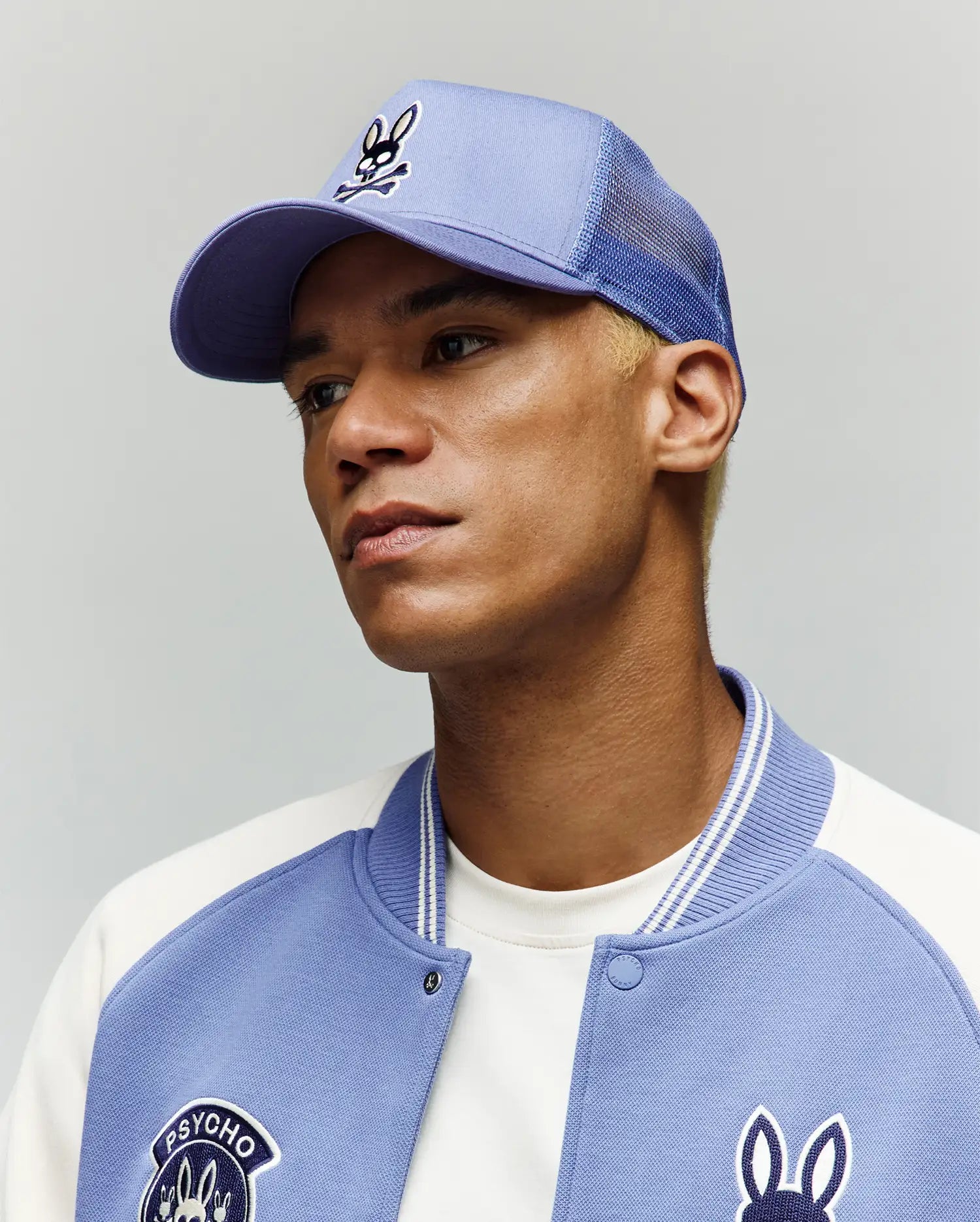 A person with short blond hair wears the light blue Psycho Bunny MENS RYDER TRUCKER CAP (MO1500030), an adjustable snapback, and a matching varsity jacket with a bunny logo, gazing slightly upward and to the side against a plain background.