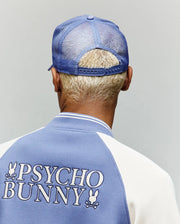A person with short blond hair is seen from behind wearing the Psycho Bunny MENS RYDER TRUCKER CAP - MO1500030 and a blue and white jacket with "PSYCHO BUNNY" and a bunny logo on the back, against a plain background.