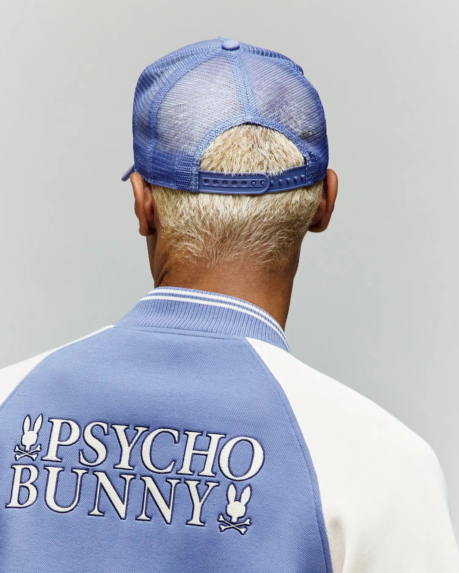 A person with short blond hair is seen from behind wearing the Psycho Bunny MENS RYDER TRUCKER CAP - MO1500030 and a blue and white jacket with "PSYCHO BUNNY" and a bunny logo on the back, against a plain background.