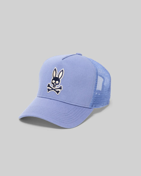 The Psycho Bunny MENS RYDER TRUCKER CAP (MO1500030) features a light blue cotton twill front, mesh back, and adjustable snapback. A bunny head above crossed bones patch adds a playful twist. Shown on a gray background.