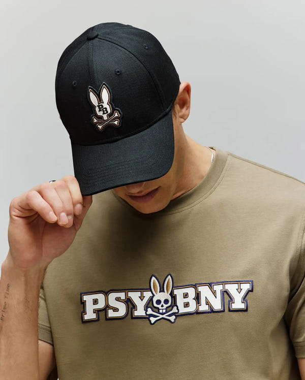 A person in a khaki shirt with "PSYBUNY" and a bunny logo looks down, touching the adjustable Psycho Bunny MENS WADE BASEBALL CAP (MO1500039) in black cotton twill, featuring an embroidered Bunny patch with crossbones.