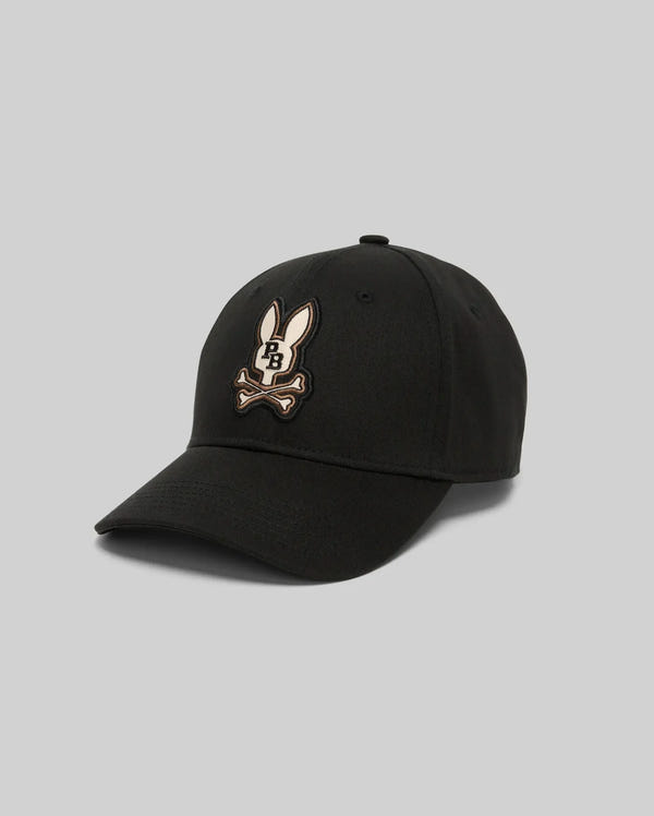 The Psycho Bunny MENS WADE BASEBALL CAP (MO1500039) is a black cotton twill cap with a curved brim, featuring an embroidered white Bunny patch above crossed bones. It has an adjustable backstrap for comfort and is shown on a plain gray background.