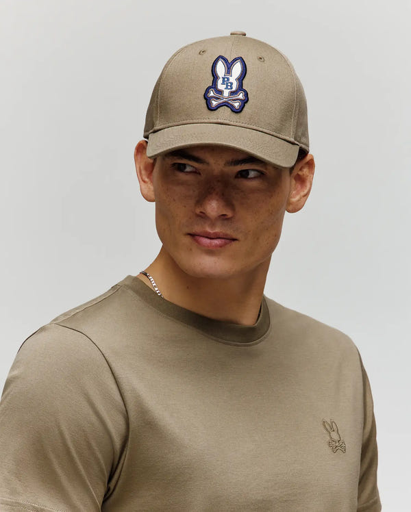 A young man wearing a khaki t-shirt and the Psycho Bunny MENS WADE BASEBALL CAP (MO1500039), featuring an embroidered Bunny patch, looks to his right against a plain light background.