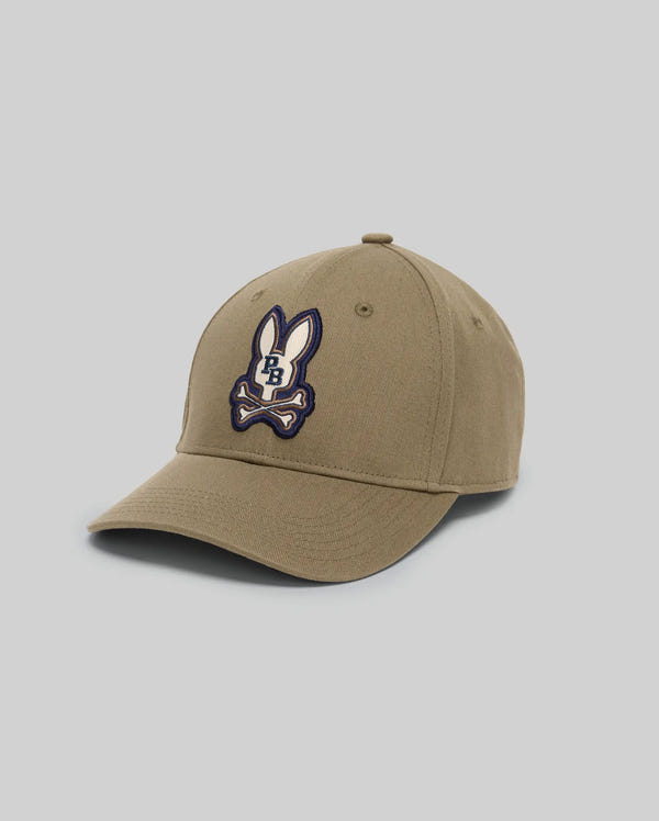 The Psycho Bunny MENS WADE BASEBALL CAP (MO1500039) is a khaki cotton twill cap with an embroidered Bunny patch, crossed bones, "PB" lettering on the front, and an adjustable backstrap, shown against a light gray background.