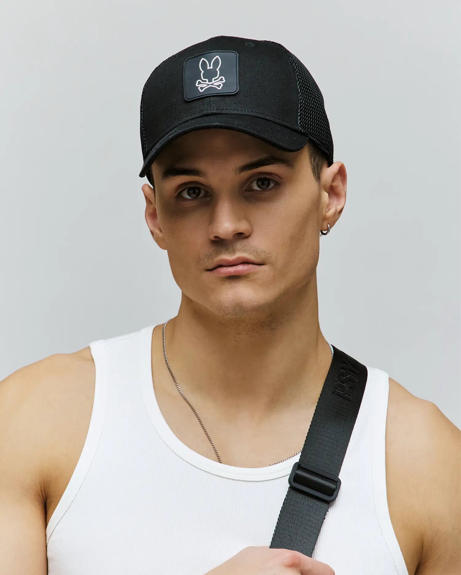 A young man wears the Psycho Bunny MENS DARIO TRUCKER CAP (MO1500044) featuring a bunny and crossbones logo, a white ribbed tank top, and a black strap across his chest, standing against a light gray background.