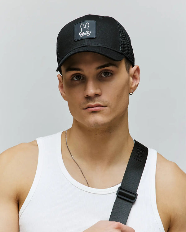 A young man wears the Psycho Bunny MENS DARIO TRUCKER CAP (MO1500044) featuring a bunny and crossbones logo, a white ribbed tank top, and a black strap across his chest, standing against a light gray background.