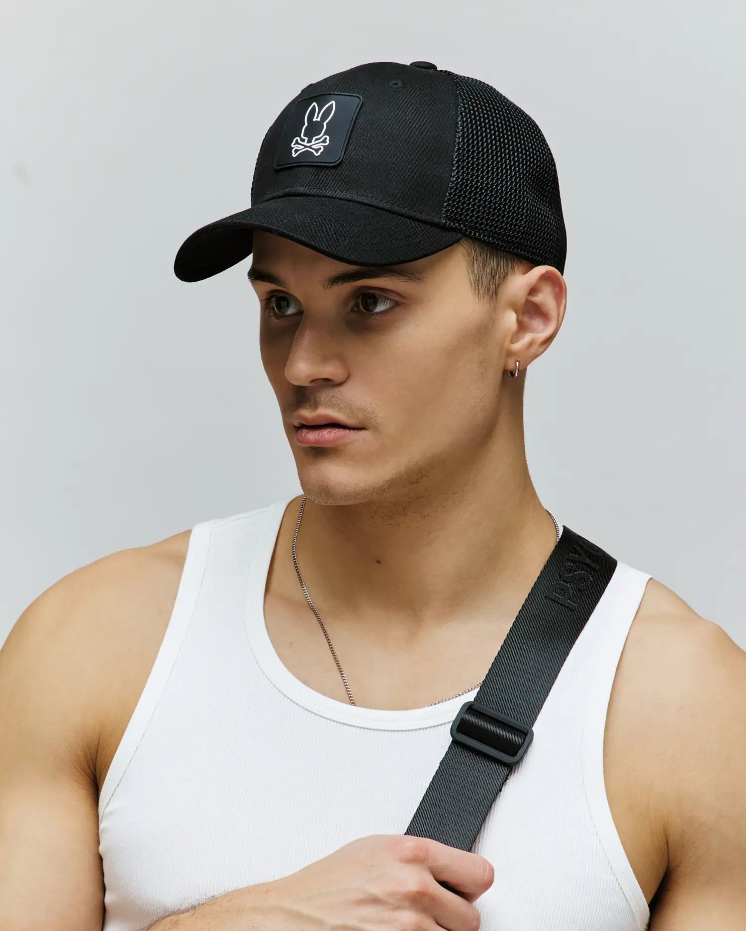 A young man stands against a plain gray background wearing a Psycho Bunny MENS DARIO TRUCKER CAP (MO1500044) featuring a breathable bunny logo and adjustable snap-back, paired with a white tank top and black chest strap.