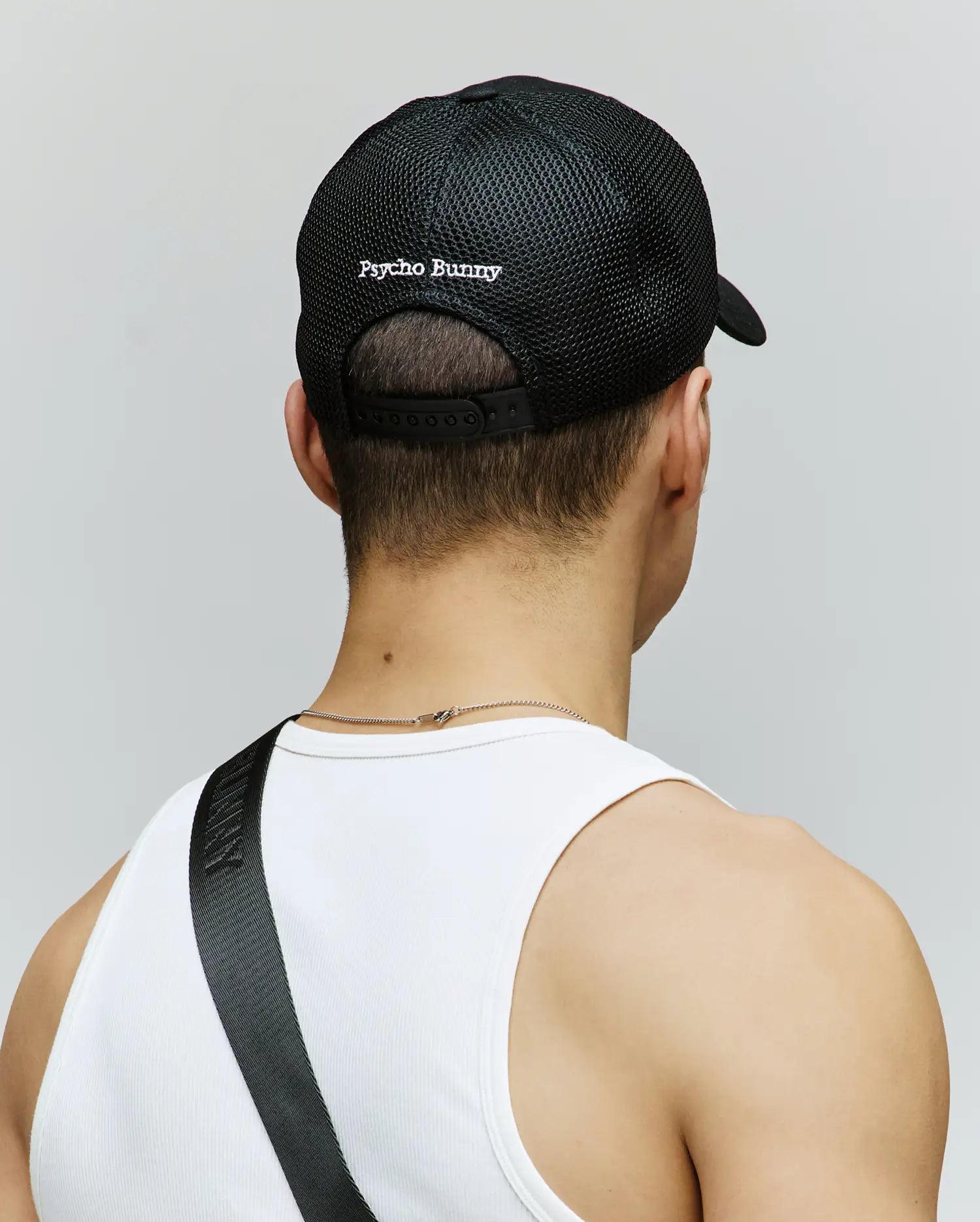 A person, seen from behind against a plain background, wears the Psycho Bunny MENS DARIO TRUCKER CAP (MO1500044) in black mesh with breathable fabric, "Psycho Bunny" text on the back, and an adjustable snap-back strap.