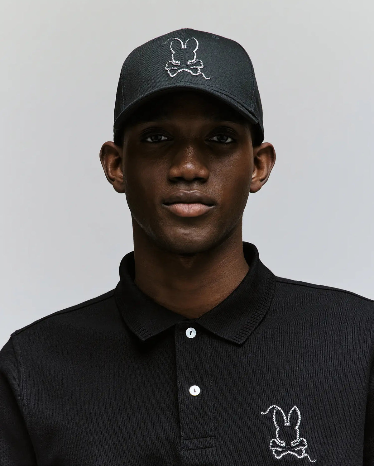 A man wears the Psycho Bunny MENS CYRUS BASEBALL CAP (MO1500045) and a black polo, both featuring the iconic white stitched bunny logo, standing against a light gray background for a blend of casual charm and modern style.