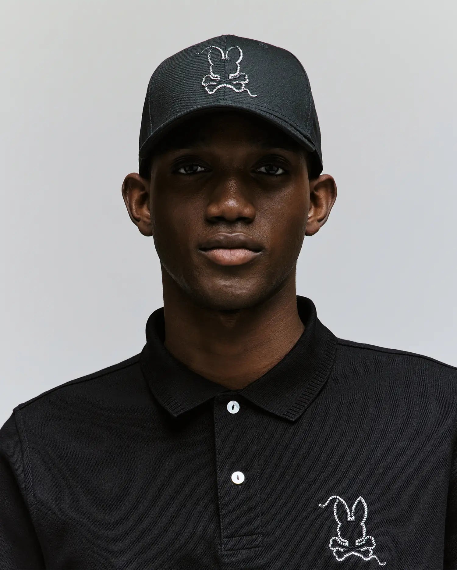 A man wears the Psycho Bunny MENS CYRUS BASEBALL CAP (MO1500045) and a black polo, both featuring the iconic white stitched bunny logo, standing against a light gray background for a blend of casual charm and modern style.