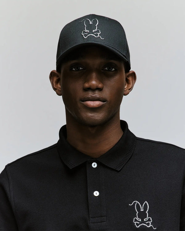 A man wears the Psycho Bunny MENS CYRUS BASEBALL CAP (MO1500045) and a black polo, both featuring the iconic white stitched bunny logo, standing against a light gray background for a blend of casual charm and modern style.