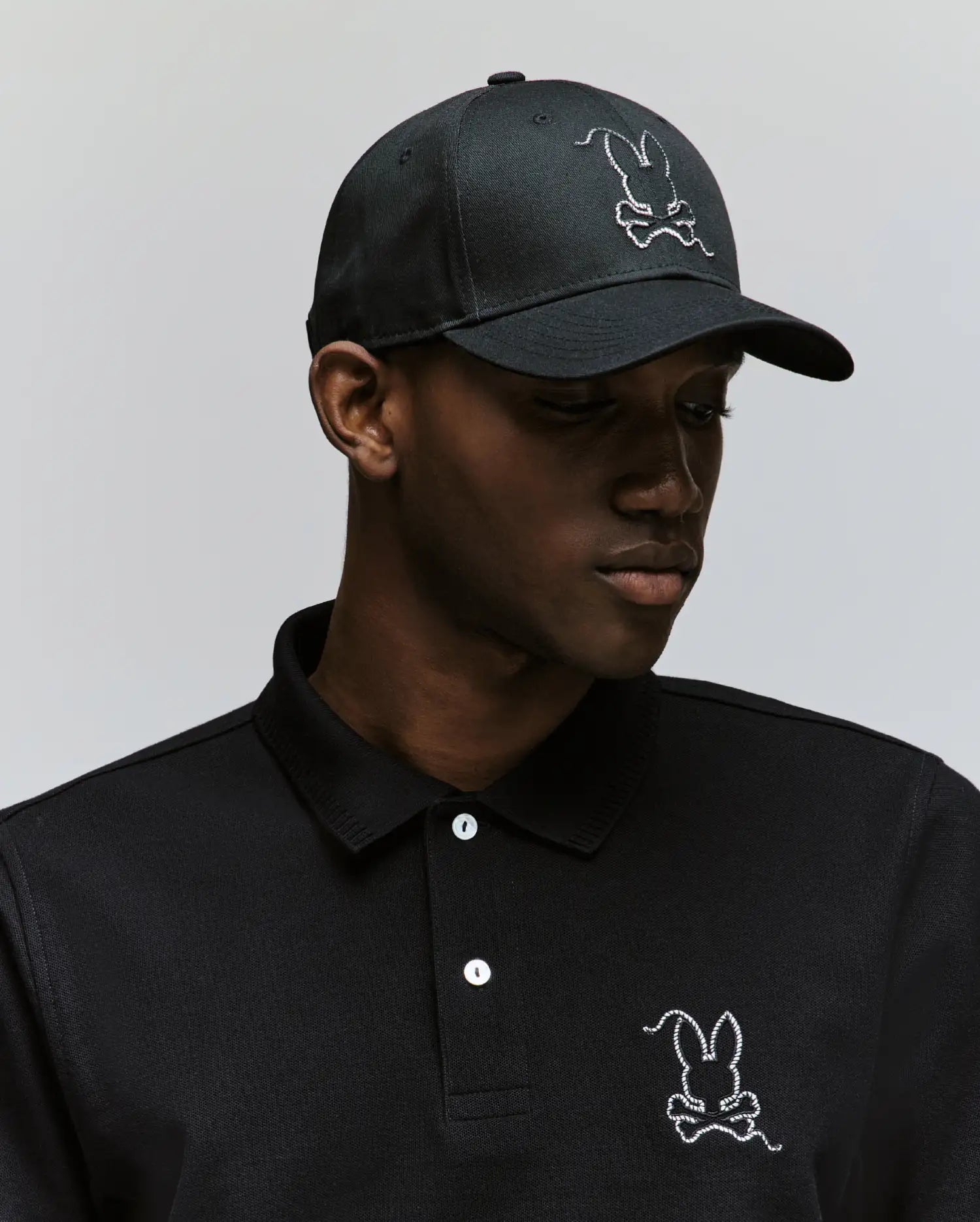 Wearing the MENS CYRUS BASEBALL CAP by Psycho Bunny and a matching black polo, each with the iconic white embroidered bunny logo, a person channels classic style while looking down against a plain background.