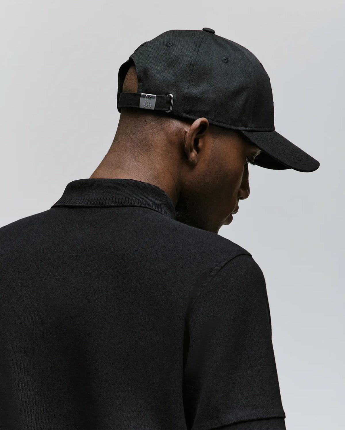 A man wearing the Psycho Bunny MENS CYRUS BASEBALL CAP (MO1500045) and a black collared shirt is seen from behind against a plain light background, giving the scene a casual, nautical vibe.