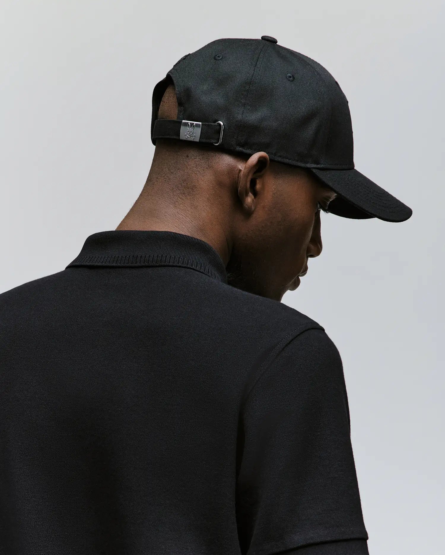 A man wearing the Psycho Bunny MENS CYRUS BASEBALL CAP (MO1500045) and a black collared shirt is seen from behind against a plain light background, giving the scene a casual, nautical vibe.