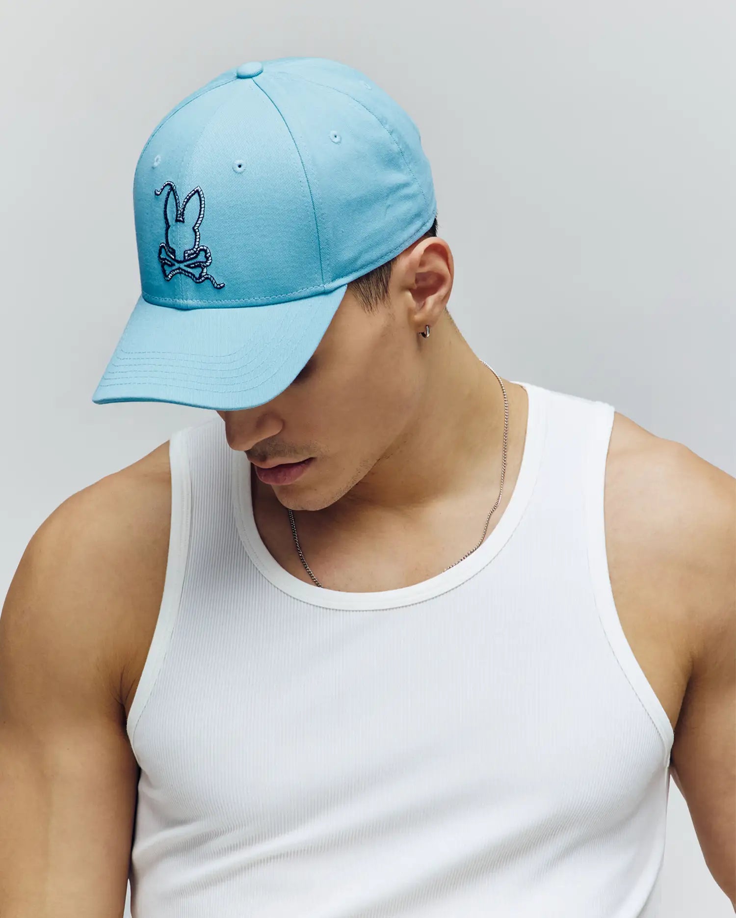 A person captures coastal charm wearing a white ribbed tank and the Psycho Bunny MENS CYRUS BASEBALL CAP - MO1500045 in light blue, featuring the bunny logo, while looking down against a plain light background.