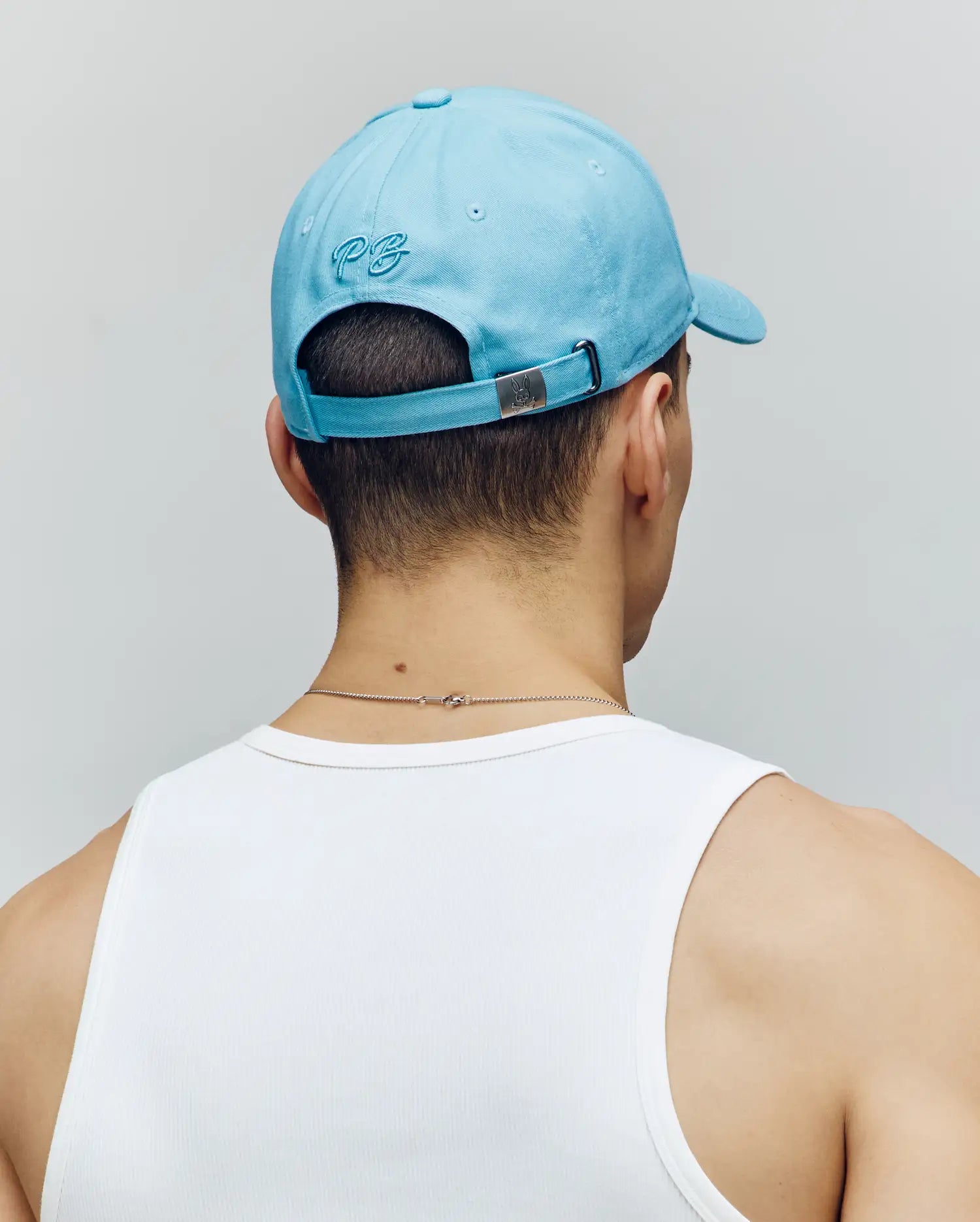 A person channels coastal charm in the Psycho Bunny Men's Cyrus Baseball Cap (MO1500045) in light blue, paired with a white tank top and thin necklace. The cap’s embroidered logo adds a nautical accent to this effortless look, seen from behind against a plain backdrop.