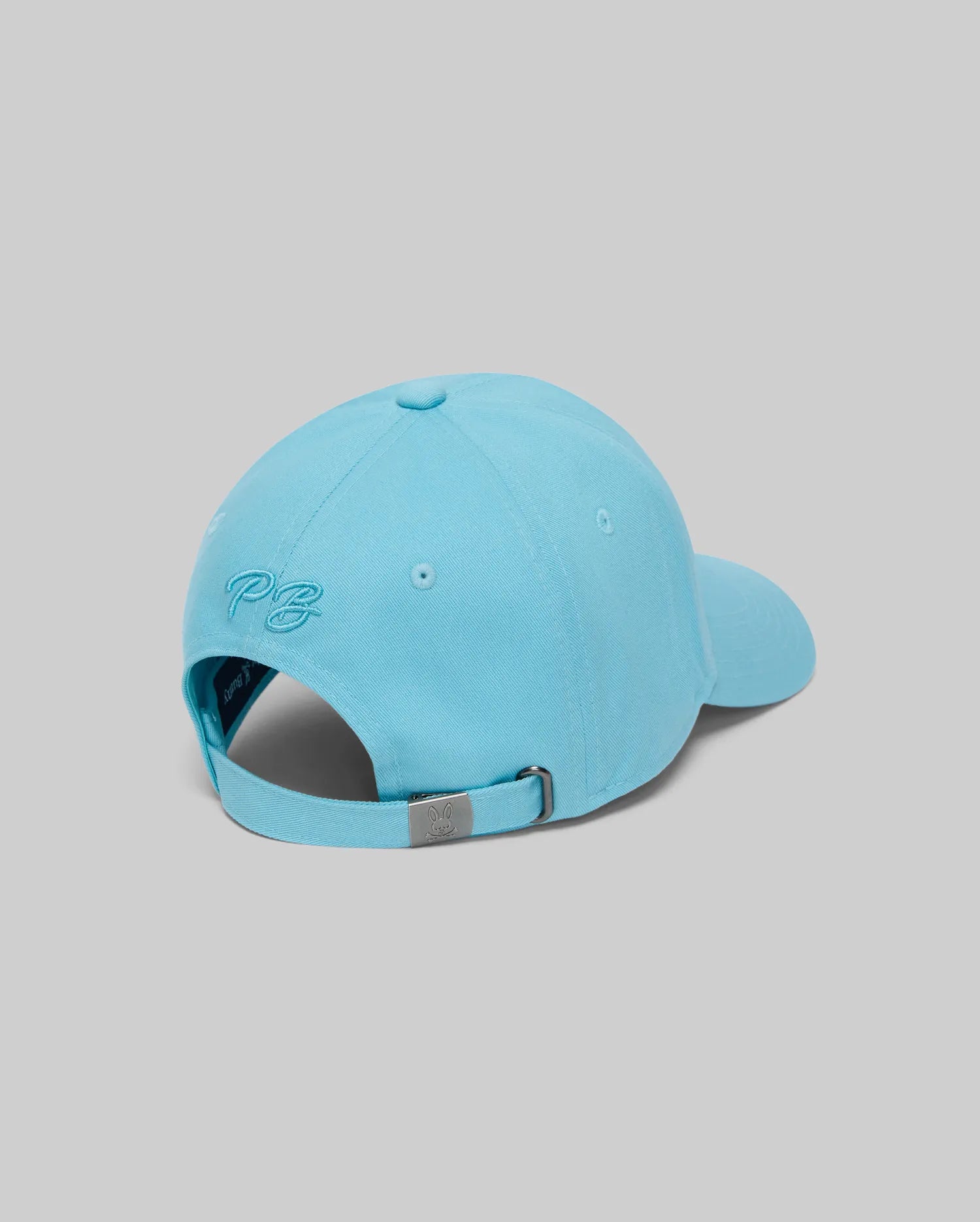 The Psycho Bunny MENS CYRUS BASEBALL CAP (MO1500045) in light blue showcases coastal and nautical style, with "PG" embroidery above an adjustable strap and metallic clasp, pictured from the back and side against a gray background.