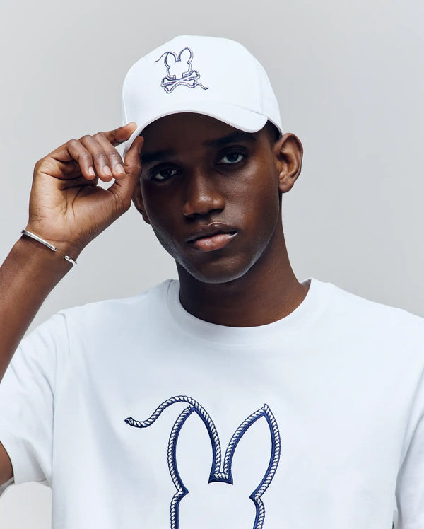 A man in the Psycho Bunny MENS CYRUS BASEBALL CAP (MO1500045), featuring the signature bunny and crossbones logo, touches his cap’s brim. He pairs it with a matching white tee, exuding subtle nautical style against a light gray background.