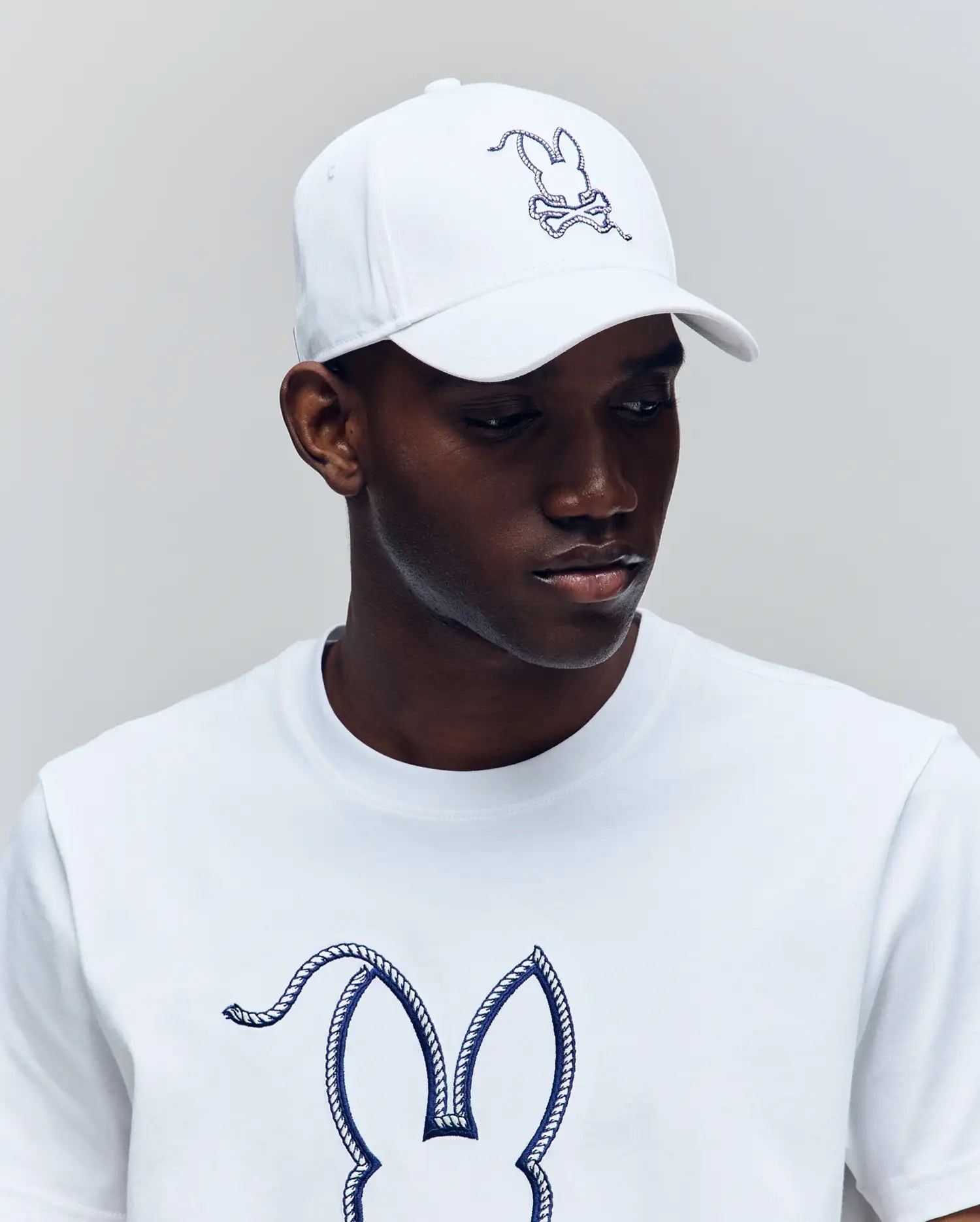A man wears the Psycho Bunny MENS CYRUS BASEBALL CAP (MO1500045) and a matching T-shirt, both with the brand’s iconic outlined bunny and crossed bones, looking down neutrally against a plain background.