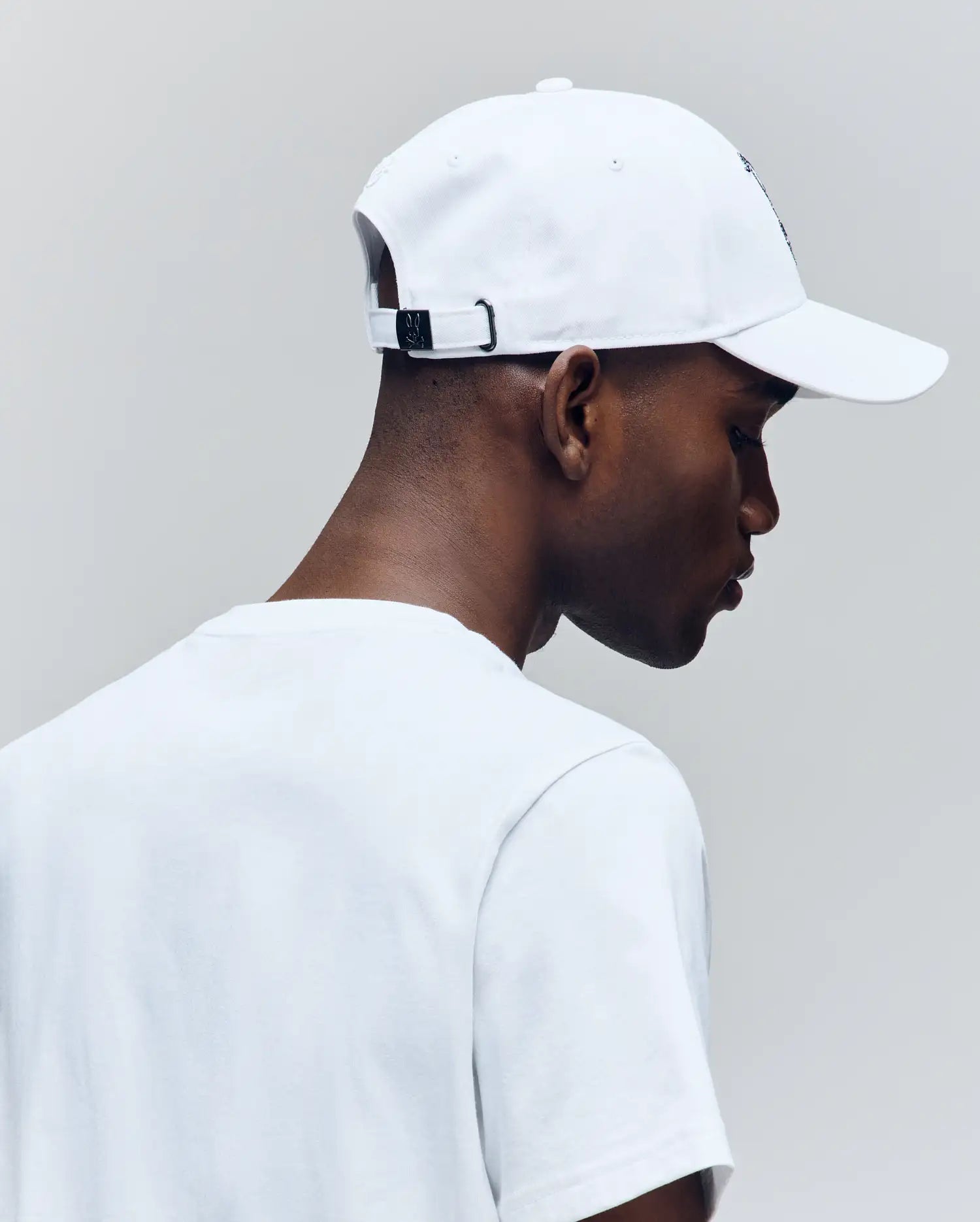 A person is seen from behind and in profile wearing the Psycho Bunny MENS CYRUS BASEBALL CAP (MO1500045) and a white t-shirt, looking down against a light grey background—evoking effortless coastal charm.