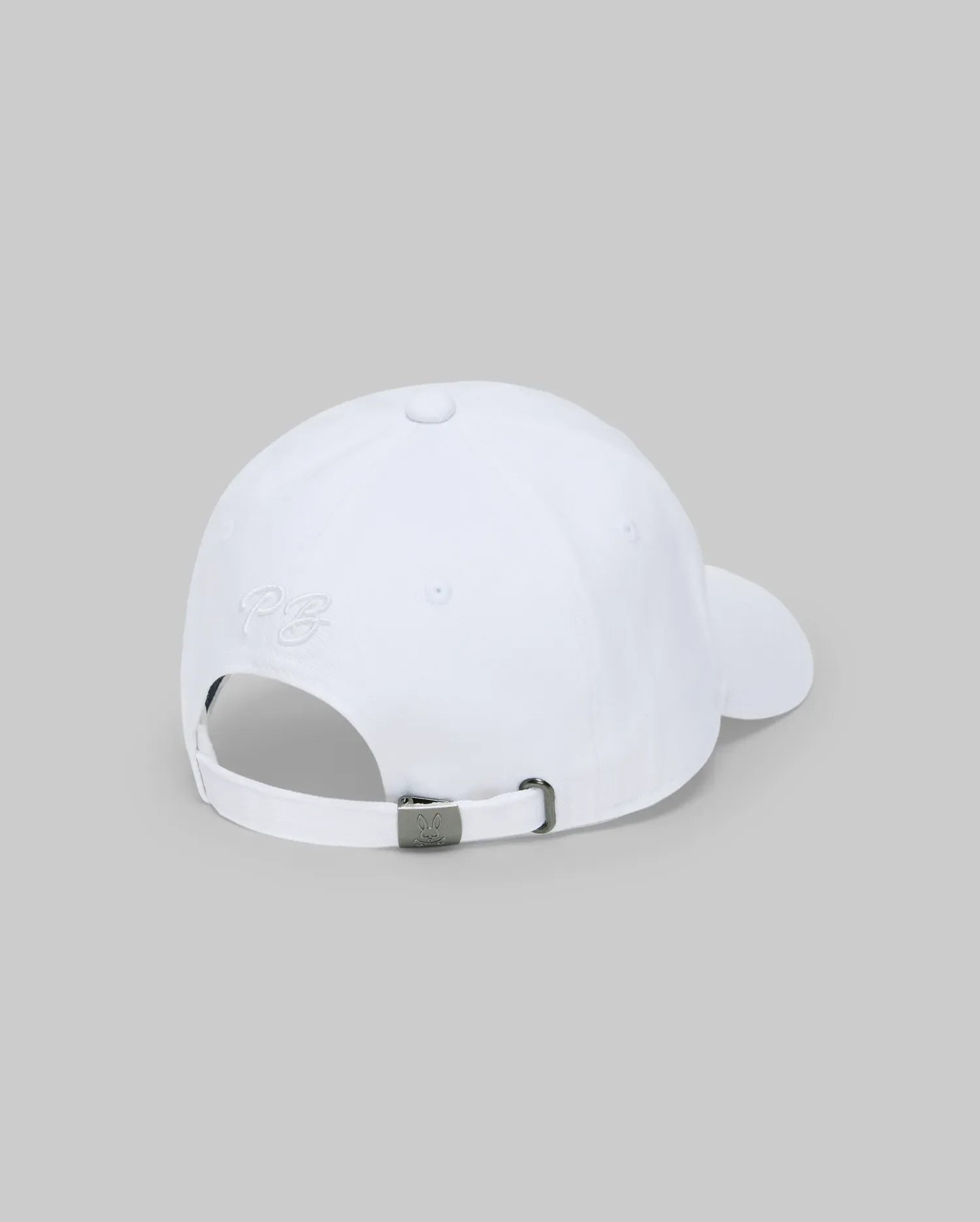 The Psycho Bunny MENS CYRUS BASEBALL CAP (MO1500045) is displayed from the back, showcasing an adjustable strap with a metal clasp and subtle side embroidery. Its white color and light gray background enhance its nautical, coastal appeal.