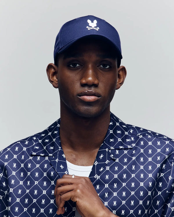 A man wearing the Psycho Bunny MENS FORREST SPORT CAP (MO1500046), featuring a navy blue color, adjustable strap, and white logo, pairs it with a patterned navy shirt while looking at the camera, holding his shirt collar.
