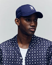 A man wears the Psycho Bunny MENS FORREST SPORT CAP (MO1500046) in navy, featuring a white bunny and crossbones logo and adjustable strap, paired with a matching navy patterned Psycho Bunny shirt and white undershirt against a plain background.