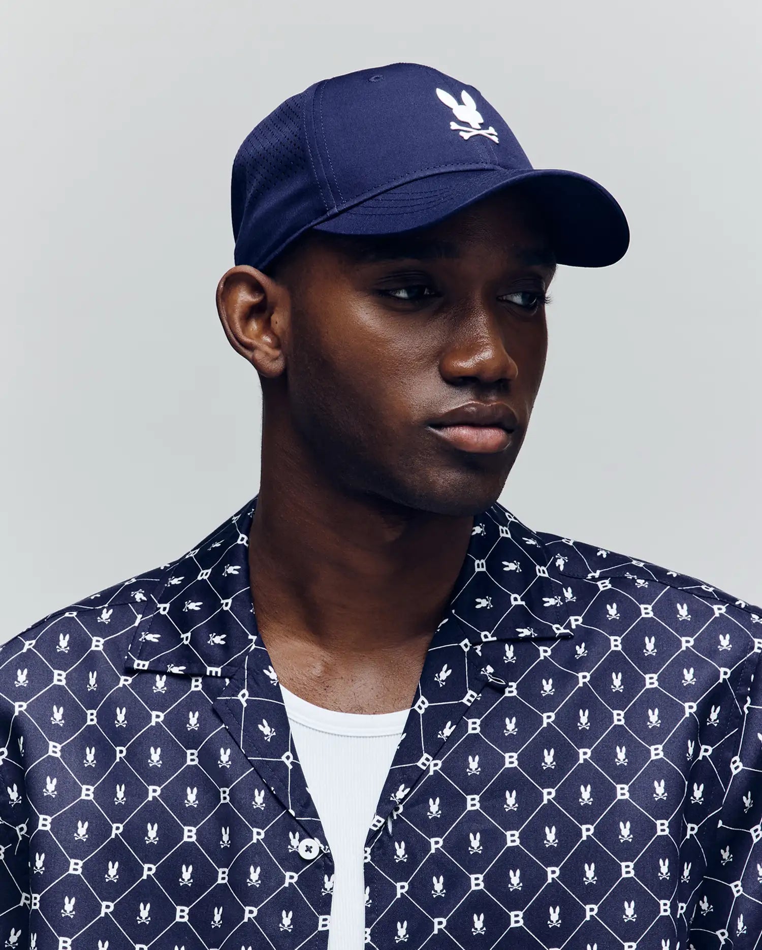 A man wears the Psycho Bunny MENS FORREST SPORT CAP (MO1500046) in navy, featuring a white bunny and crossbones logo and adjustable strap, paired with a matching navy patterned Psycho Bunny shirt and white undershirt against a plain background.