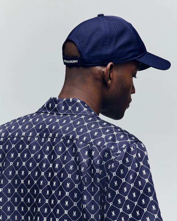 A man is seen from behind against a plain light background, wearing the Psycho Bunny MENS FORREST SPORT CAP (MO1500046), a navy blue adjustable cap, and a patterned shirt with small bunny logos.