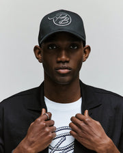 A person wears a Psycho Bunny MENS CYRUS PB BASEBALL CAP - MO1500047, a black jacket, and a white Cyrus tee with a rope-style logo. Facing forward and gripping their jacket collar, they embody a seafaring vibe.