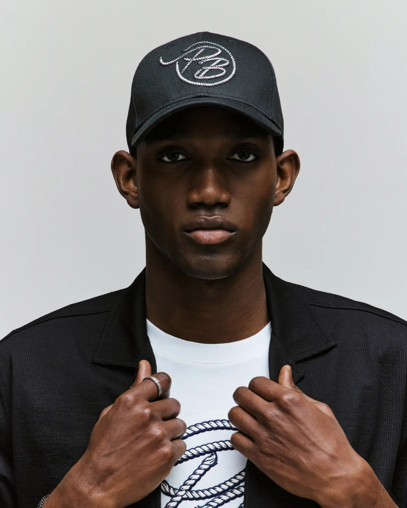 A person wears a Psycho Bunny MENS CYRUS PB BASEBALL CAP - MO1500047, a black jacket, and a white Cyrus tee with a rope-style logo. Facing forward and gripping their jacket collar, they embody a seafaring vibe.
