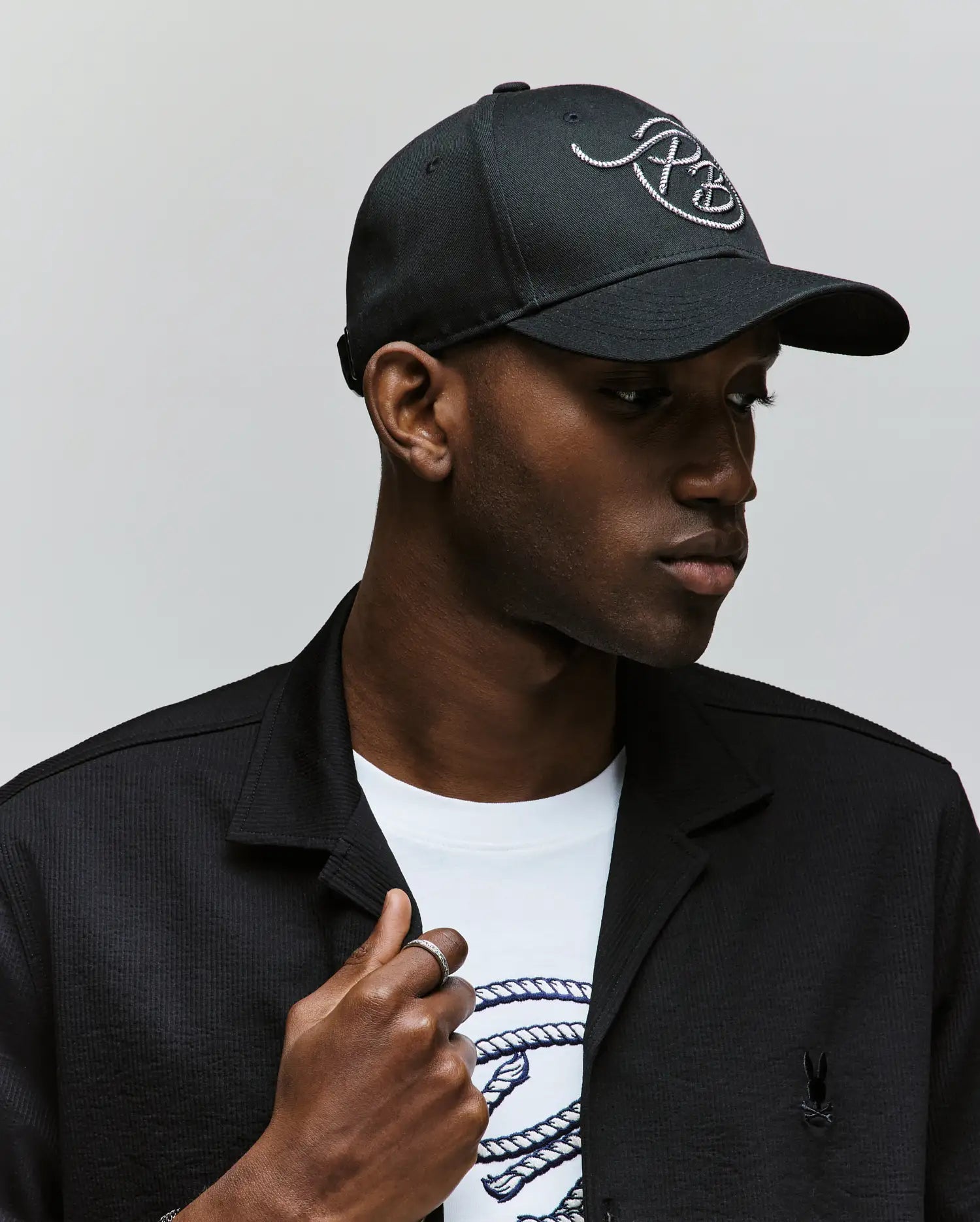 A man in a Psycho Bunny MENS CYRUS PB BASEBALL CAP (MO1500047), black jacket, and white printed Cyrus tee looks left against a plain light background.