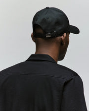 A person is seen from behind wearing the Psycho Bunny MENS CYRUS PB BASEBALL CAP - MO1500047 in black and a textured black jacket with a rope-style logo, standing against a plain light gray background.