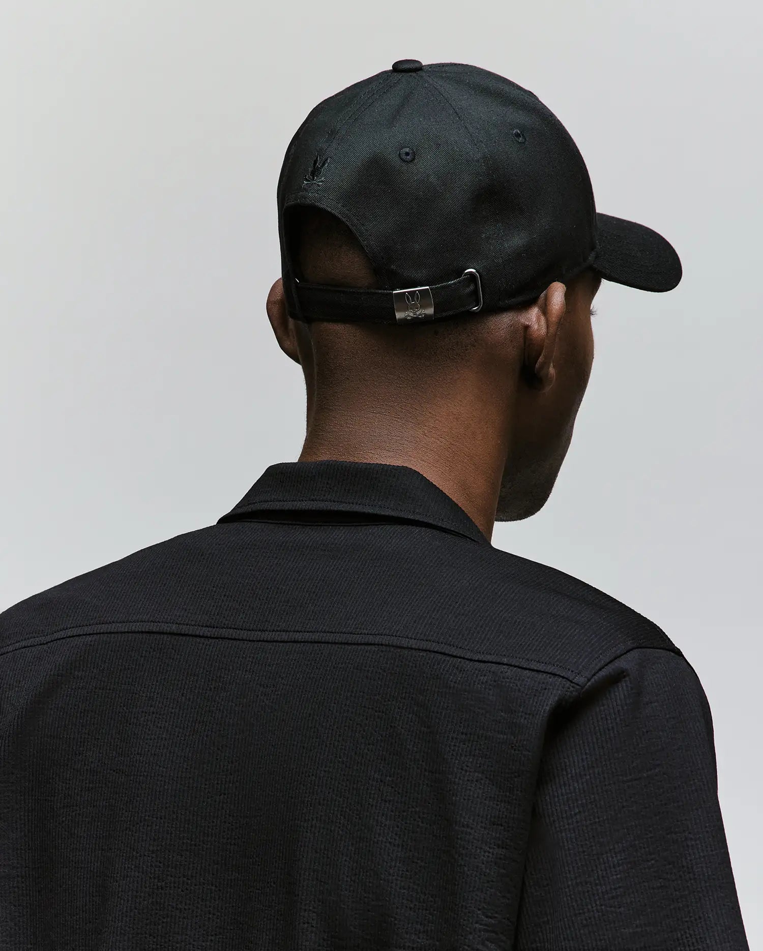 A person is seen from behind wearing the Psycho Bunny MENS CYRUS PB BASEBALL CAP - MO1500047 in black and a textured black jacket with a rope-style logo, standing against a plain light gray background.