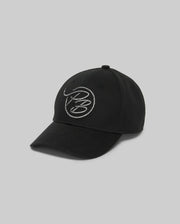 The Psycho Bunny MENS CYRUS PB BASEBALL CAP - MO1500047 is a black baseball cap with a white circular rope-style abstract embroidered logo on the front, shown against a plain grey background.