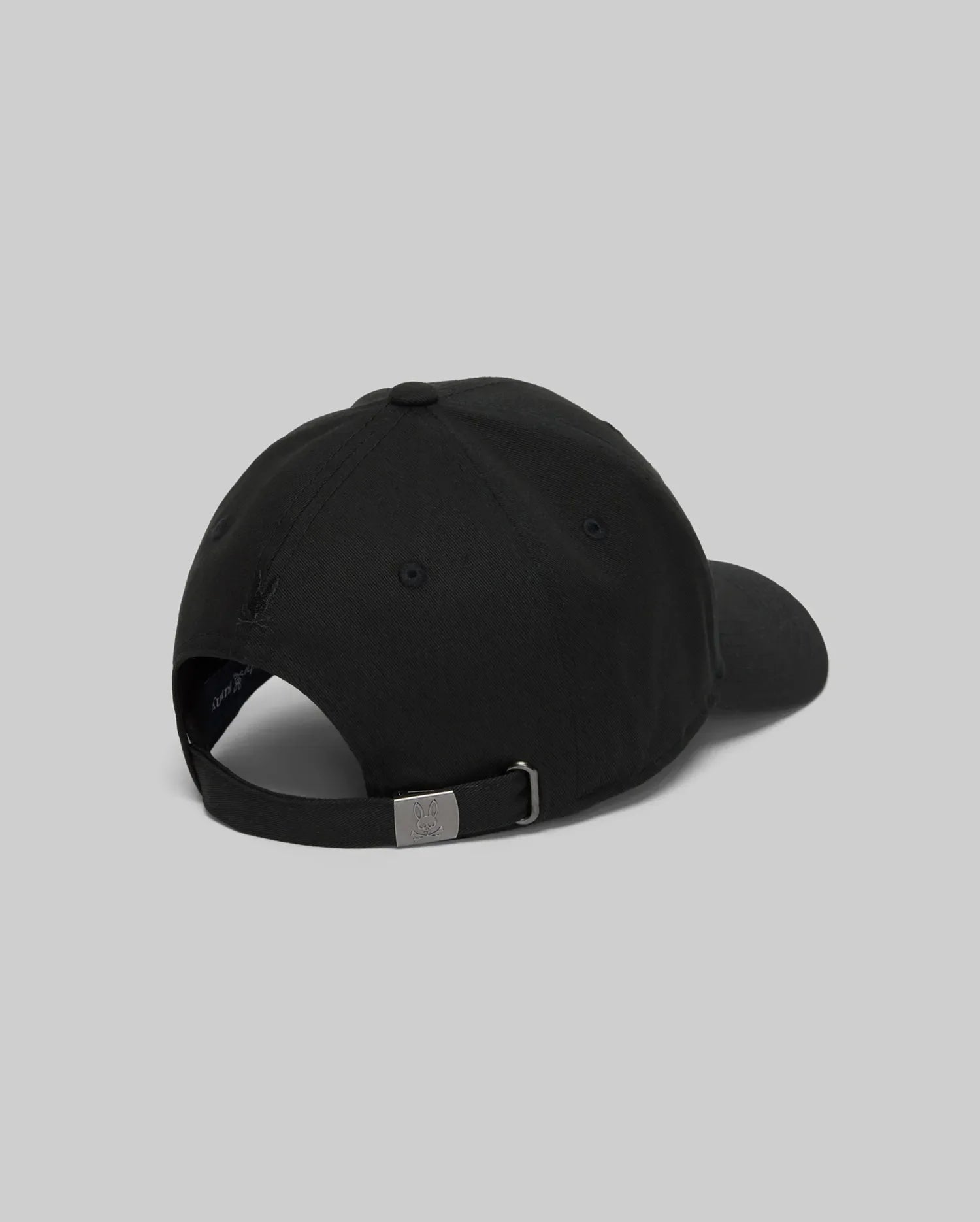 The Psycho Bunny MENS CYRUS PB BASEBALL CAP - MO1500047 in black is shown from the back, featuring a curved brim, adjustable metal-buckle strap, and a subtle rope-style logo graphic against a plain gray background.