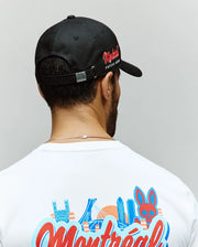A man is seen from behind wearing a Psycho Bunny MENS MONTREAL BASEBALL CAP - MO1500048 in black with red side text, paired with a white 100% cotton tee featuring colorful city buildings and an embroidered Bunny graphic.