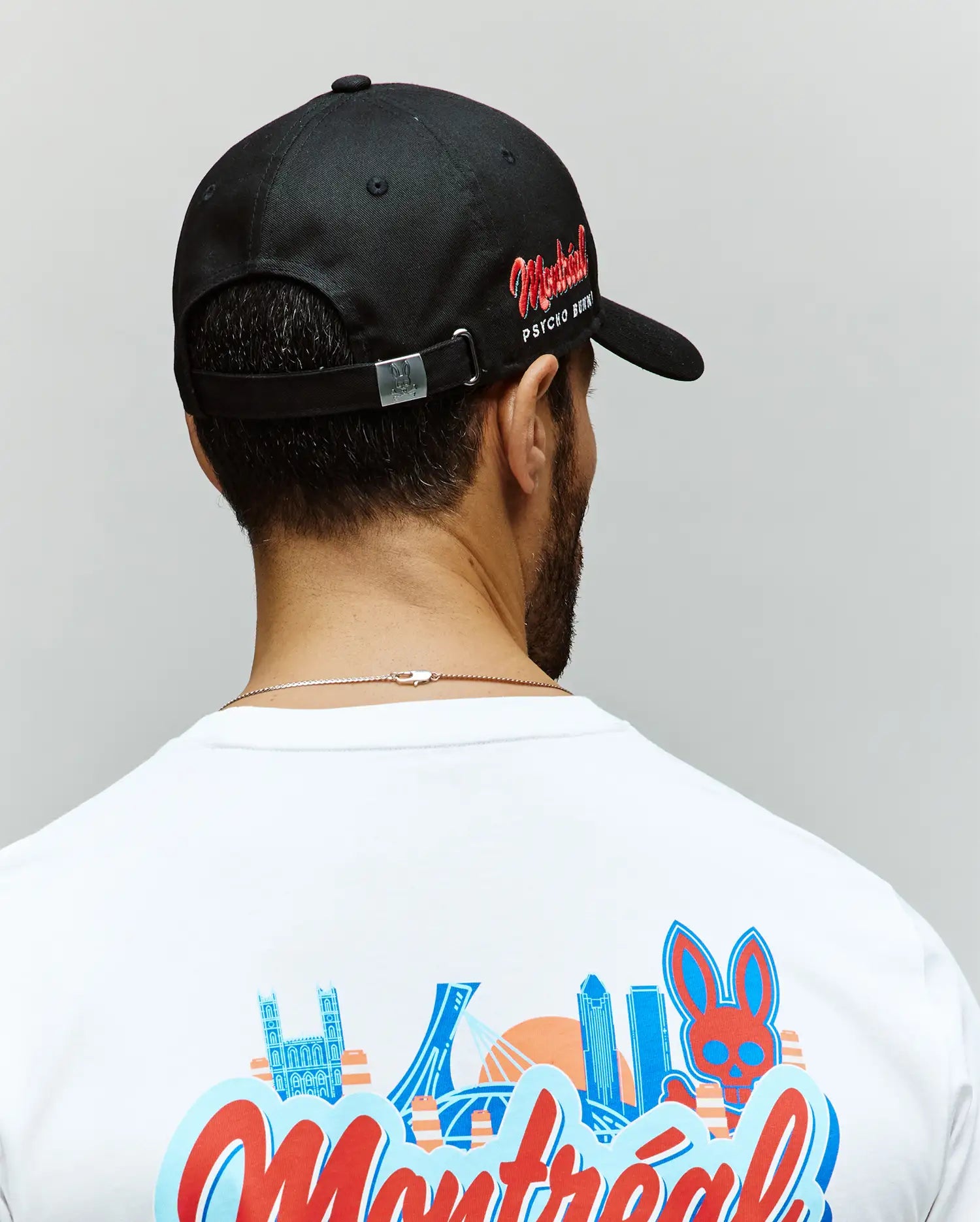 A man is seen from behind wearing a Psycho Bunny MENS MONTREAL BASEBALL CAP - MO1500048 in black with red side text, paired with a white 100% cotton tee featuring colorful city buildings and an embroidered Bunny graphic.