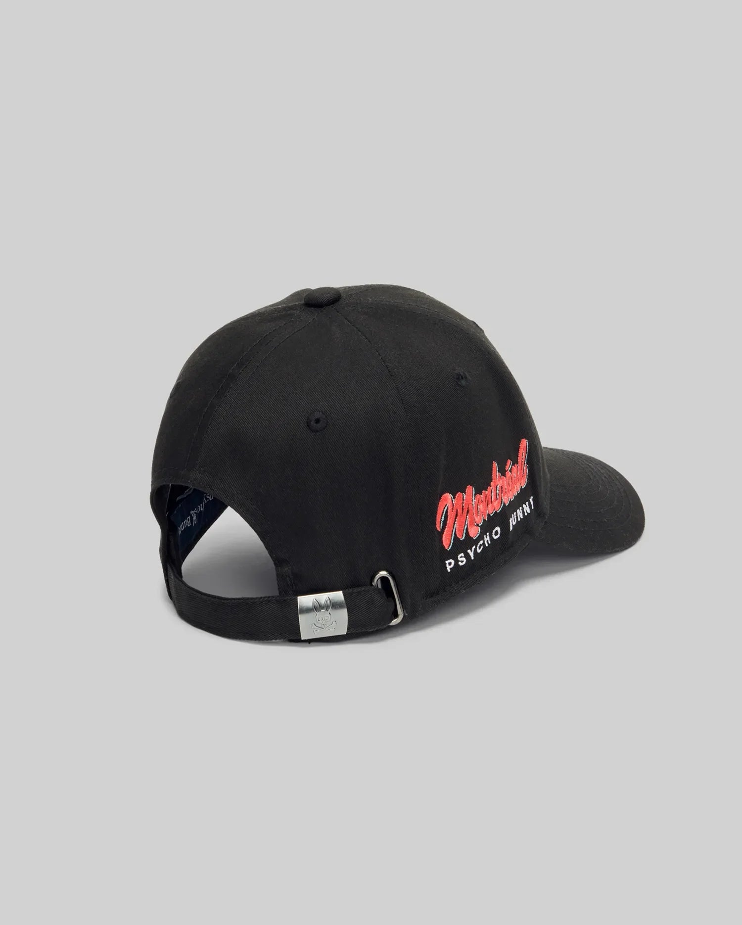 A rear view of the Psycho Bunny MENS MONTREAL BASEBALL CAP (MO1500048), black and 100% cotton, with an adjustable metal-clip strap and red and white "Montreal PSYCHO PRINT" embroidery on the side. Light gray background.