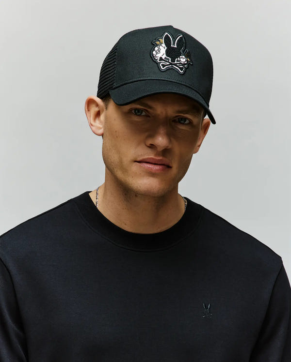 A man wearing the Psycho Bunny MENS AUGUSTE TRUCKER CAP - MO1500050 and a black crew-neck sweatshirt stands against a plain light gray background.