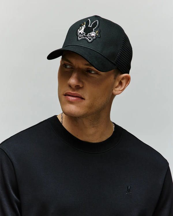 A man in a Psycho Bunny MENS AUGUSTE TRUCKER CAP (MO1500050) and a black sweatshirt looks slightly to the side against a plain, light background.