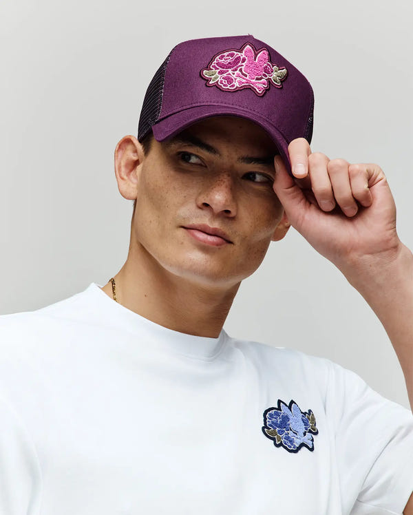 Wearing a white shirt with a blue flower patch, a person holds the brim of the Psycho Bunny MENS AUGUSTE TRUCKER CAP (MO1500050), which features a pink floral chenille patch, looking slightly to the side against a neutral background.