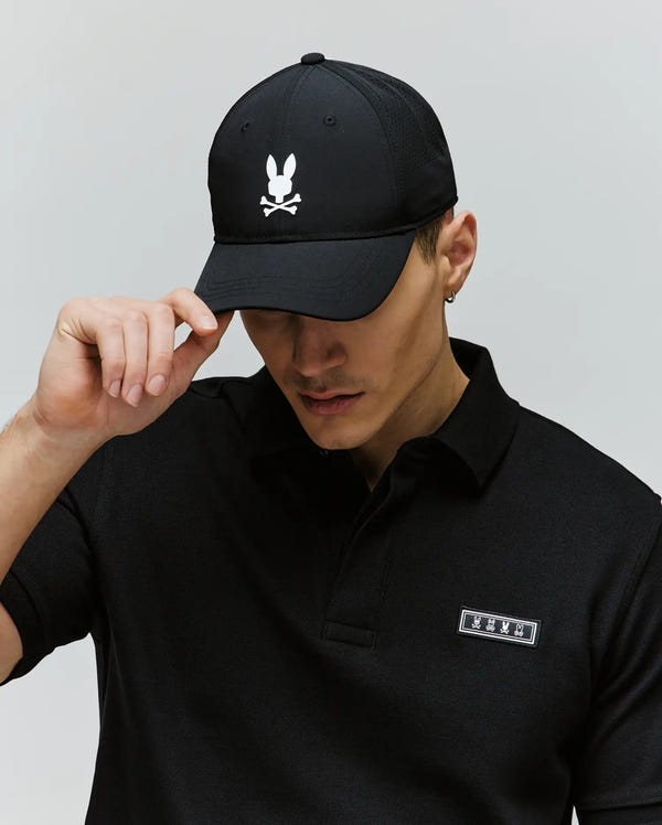 A person wearing a black polo and the Psycho Bunny MENS FORREST SPORT CAP (MO1500051), a black cap with a white bunny skull-and-crossbones logo and adjustable strap, holds the brim and looks downward against a plain background.