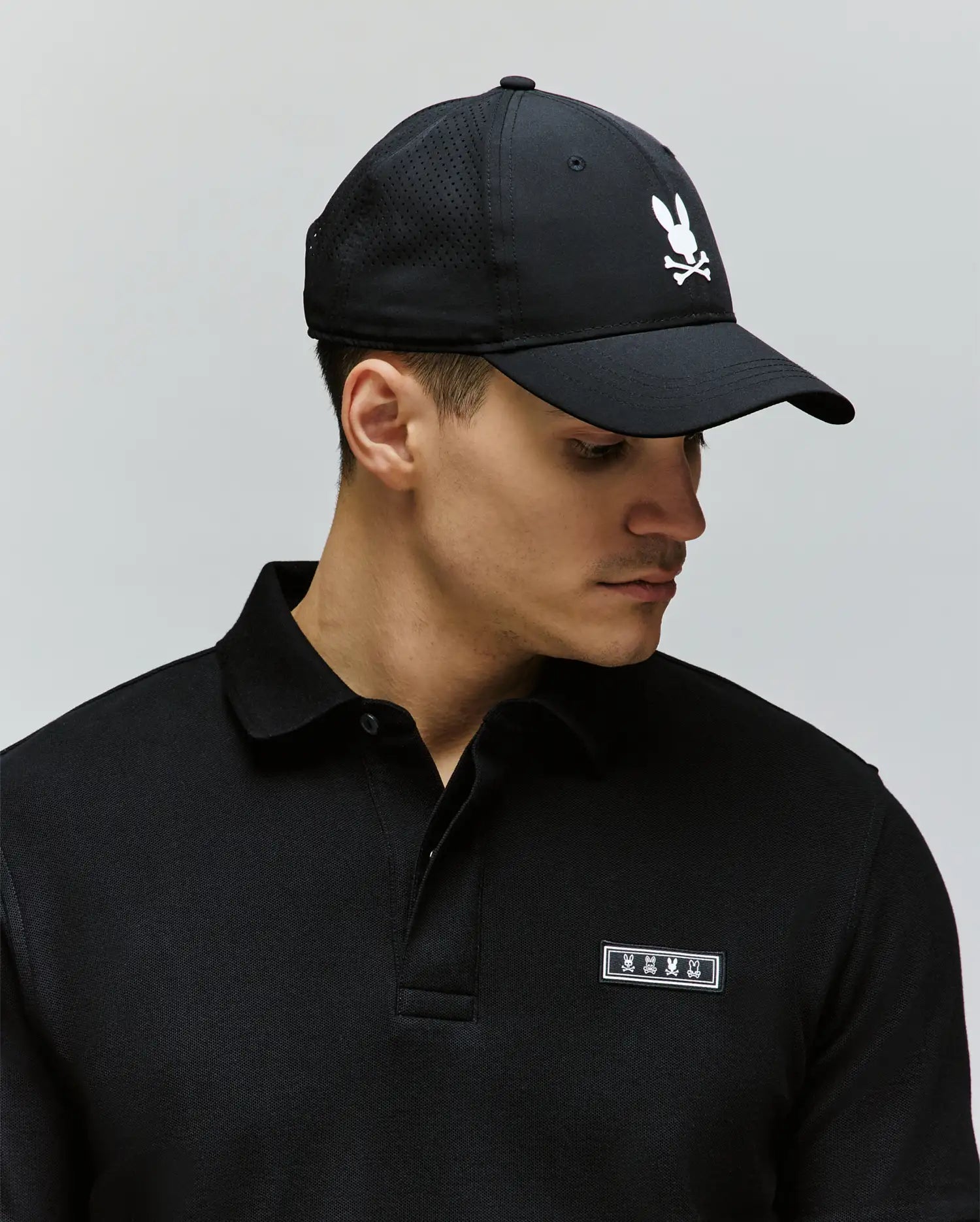 A man wearing a black collared shirt and the Psycho Bunny MENS FORREST SPORT CAP (MO1500051), featuring an adjustable strap and a white bunny-and-crossbones logo, looks down to the side against a plain background.