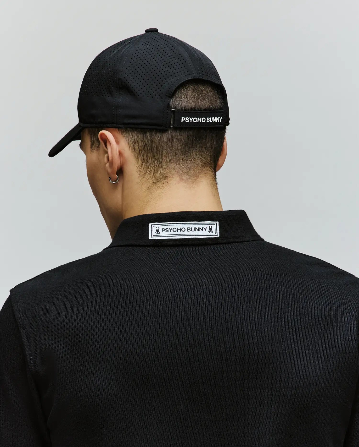 A person is seen from the back against a plain light background, wearing the Psycho Bunny MENS FORREST SPORT CAP (MO1500051) and a black polo shirt, both featuring Psycho Bunny branding.