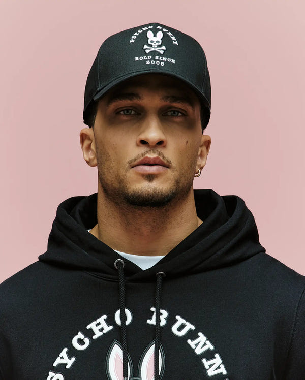 A man wearing the Psycho Bunny MENS ARCHIVE BASEBALL CAP - MO1500002 with a skull and crossbones logo and a black hoodie with an embroidered bunny and "Psycho Bunny" text stands against a plain pink background.