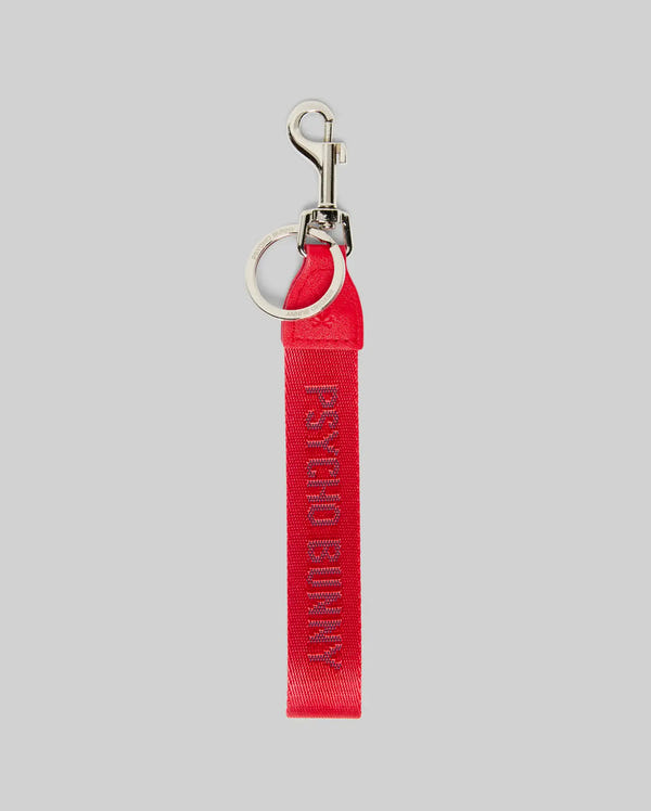 The PSYCHO BUNNY WOVEN TAPE KEYCHAIN - MO1700001 by Psycho Bunny is a red fabric keychain with a durable design, silver metal ring and clasp, and "PSYCHO BUNNY" embroidered in pink on the ribbon, shown on a light gray background.