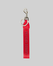The PSYCHO BUNNY WOVEN TAPE KEYCHAIN - MO1700001 by Psycho Bunny is a red fabric keychain with a durable design, silver metal ring and clasp, and "PSYCHO BUNNY" embroidered in pink on the ribbon, shown on a light gray background.
