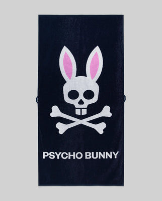 The Psycho Bunny MENS CLASSIC BUNNY TOWEL (MO1700004) is a dark towel with a white skull and crossbones topped by pink bunny ears above bold "PSYCHO BUNNY" lettering.